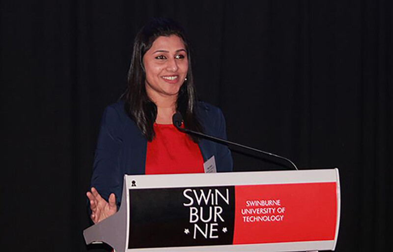 Nisa Salim stands at a podium which reads "Swinburne". She wears a red shirt with a black cardigan. She is smiling broadly.