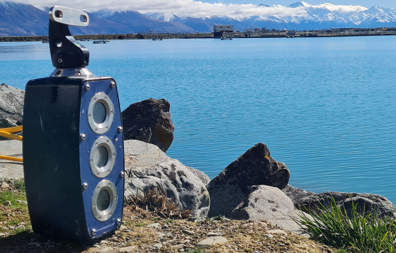 An Ace Aquatec A-Biomass camera sitting on the shoreline of an alpine lake with bright blue water.