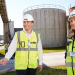 Two people wearing construction helmets and Enifer-branded high visibility vests in front of large storage tanks.