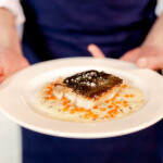 A chef in a blue apron holding a plate with a piece of cooked sablefish on it
