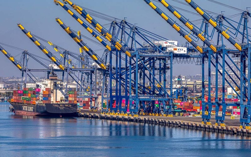 Blue and yellow cranes suspended over the port of Los Angeles, where a blue container ship waits, stacked high with multicolored containers.