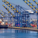Blue and yellow cranes suspended over the port of Los Angeles, where a blue container ship waits, stacked high with multicolored containers.
