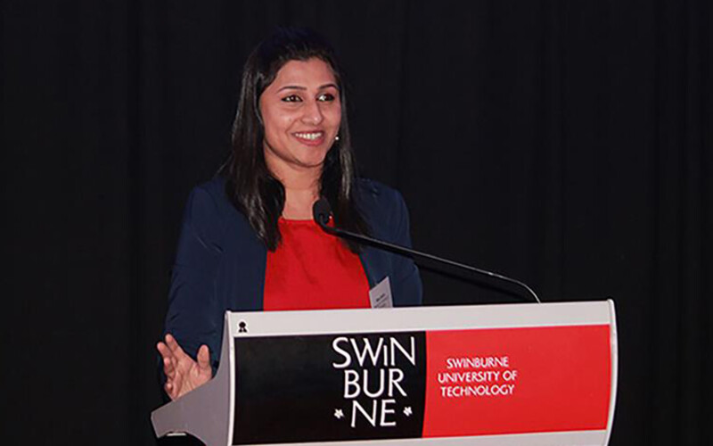 Nisa Salim stands at a podium which reads "Swinburne". She wears a red shirt with a black cardigan. She is smiling broadly.
