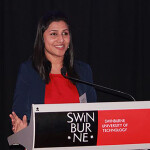 Nisa Salim stands at a podium which reads "Swinburne". She wears a red shirt with a black cardigan. She is smiling broadly.