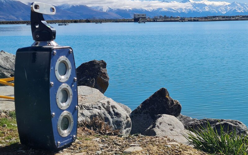 An Ace Aquatec A-Biomass camera sitting on the shoreline of an alpine lake with bright blue water.