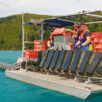 Two white men wearing protective gear stand on a floating platform surrounded by teal water and load orange baskets full of oysters into a "flipper" device.