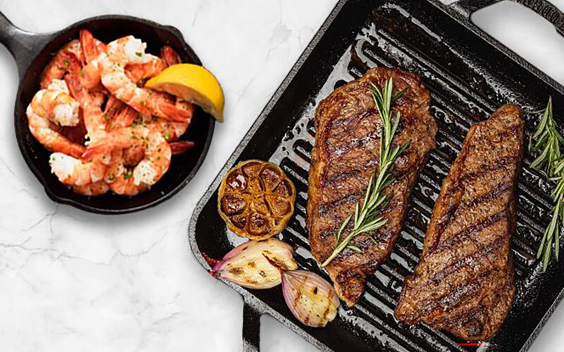 Two steaks on a griddle next to a cast iron skillet full of shrimp