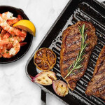 Two steaks on a griddle next to a cast iron skillet full of shrimp