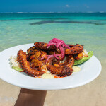 A person holding grilled octopus up to Caribbean waters